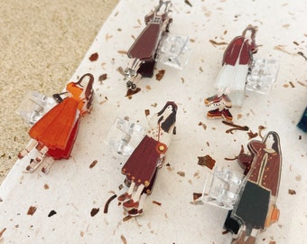 May include: Five colorful hair clips featuring cartoon women in different outfits. The clips are made of clear plastic with silver metal clasps.