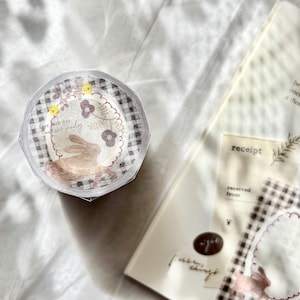 May include: A roll of decorative washi tape featuring a gingham pattern and a design with a bunny, flowers, and the text "Live each day sweet and simply". The tape is partially unwound, revealing the design.