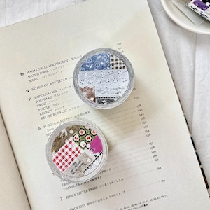 May include: Two clear, round containers filled with colourful fabric scraps sit on an open book. The containers have a patchwork design with floral, gingham, and lace patterns. A small plate with similar decorative tape rolls is in the background.