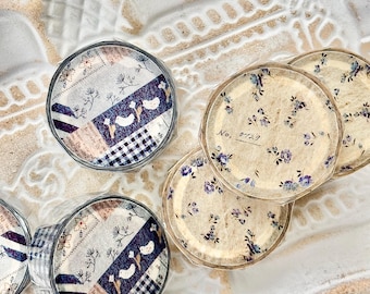 Puede incluir: Varios rollos circulares de cinta decorativa washi envueltos en plástico transparente. Dos rollos presentan un diseño de patchwork con pájaros, motivos florales y cuadros vichy azules. Tres rollos muestran un fondo crema con flores pequeñas.