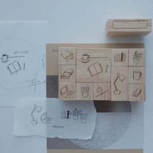 May include: A collection of light-colored wooden stamps featuring engraved line art illustrations of office and home items including a laptop, books, a desk lamp, a coffee mug, a potted plant, and a smartphone, alongside a rectangular wooden block.