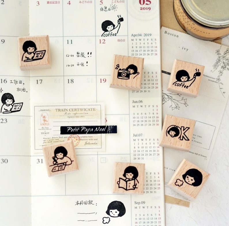 Puede incluir: Una colecci&oacute;n de peque&ntilde;os sellos cuadrados de madera con ilustraciones en tinta negra de una ni&ntilde;a con una c&aacute;mara, una taza de caf&eacute; y un port&aacute;til. Los sellos est&aacute;n colocados sobre un calendario.