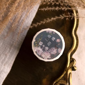 May include: A roll of washi tape featuring a floral design. The tape is decorated with purple and white flowers on a dark blue background. The text on the tape reads "1cm x 0.5m" and "霧花".