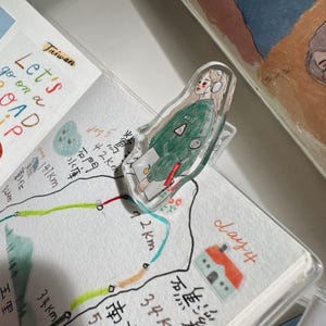 May include: A clear acrylic bookmark featuring a cartoon illustration of a person wearing headphones and a green jacket, holding a laptop and a drink. The bookmark is clipped to a notebook with a map and the text "Let's go on a ROAD TRIP".