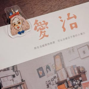 May include: A clear acrylic bookmark featuring a stylized illustration of a woman with pink hair and a blue bow. The bookmark is placed on a white page with orange Chinese characters. The page also has a printed interior scene.