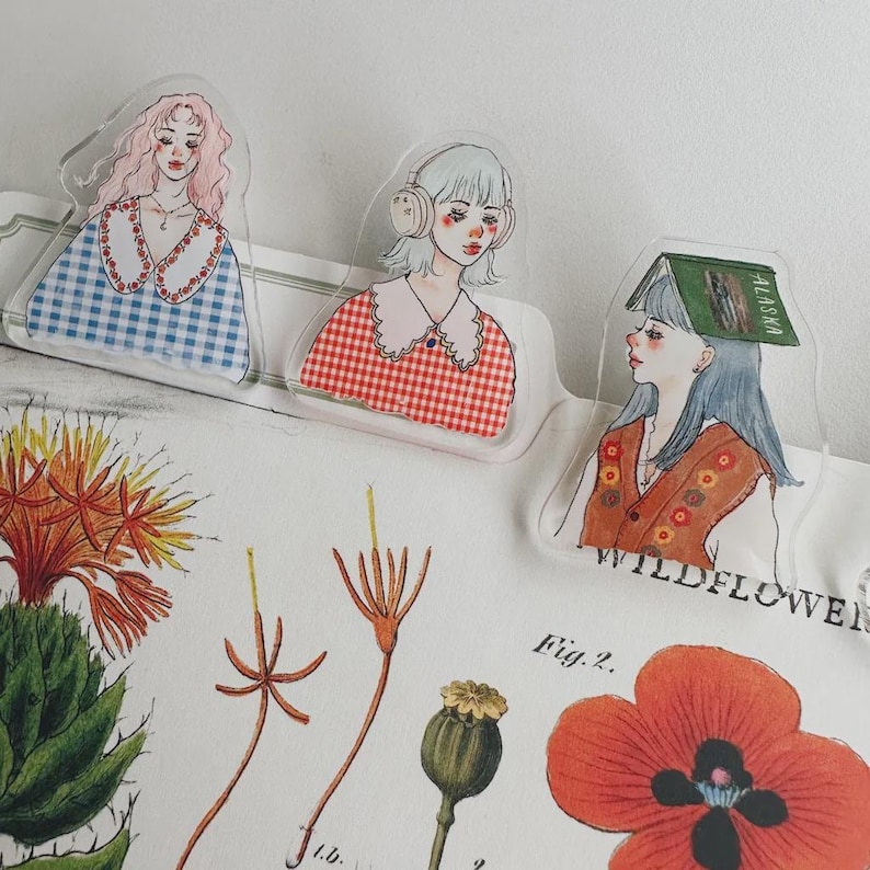 May include: Three clear acrylic bookmarks with illustrations of young women. One wears a blue gingham dress, another has headphones, and the third has a book on her head. The bookmarks are placed over a botanical illustration.