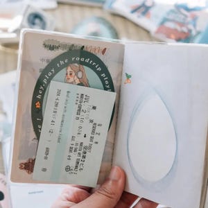 May include: A passport holder with a clear plastic cover, featuring a sticker with a cartoon illustration of a woman with headphones and the text "hey, play the roadtrip play!"  The holder also contains a train ticket with the text "JUL. 2 (10:03) (10:54) MARITA-EXPRESS 15" and "TOKYO NARITA AIR TERMINAL-3".