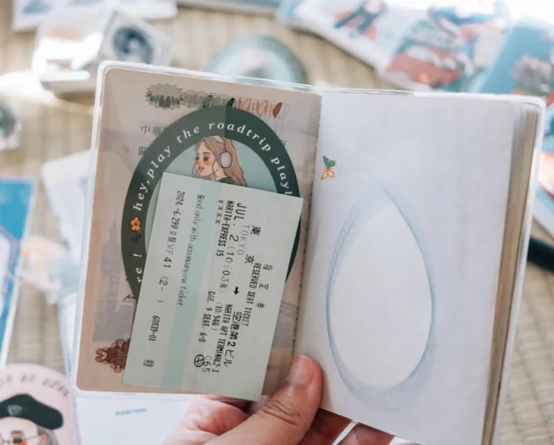 May include: A passport holder with a clear plastic cover, featuring a sticker with a cartoon illustration of a woman with headphones and the text "hey, play the roadtrip play!"  The holder also contains a train ticket with the text "JUL. 2 (10:03) (10:54) MARITA-EXPRESS 15" and "TOKYO NARITA AIR TERMINAL-3".