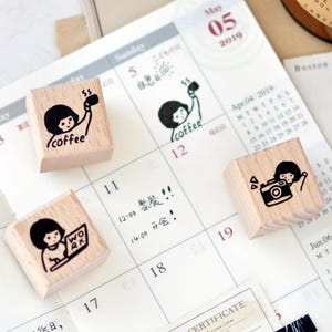 Puede incluir: Cuatro sellos de madera con ilustraciones en tinta negra sobre un calendario. Un sello dice "coffee", otro muestra a una persona con una taza de caf&eacute;, un tercero muestra a una persona con una c&aacute;mara y el &uacute;ltimo muestra a una persona trabajando en una computadora port&aacute;til.