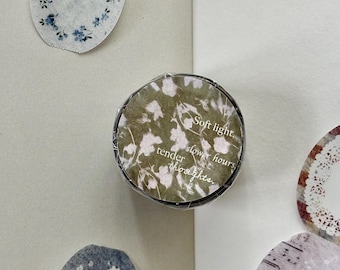 Puede incluir: Un rollo de cinta decorativa washi con fondo verde oliva, patrones florales blancos y el texto Soft light, slower hours, tender thoughts. Alrededor del rollo hay varias pegatinas de papel ovaladas con diseños florales.