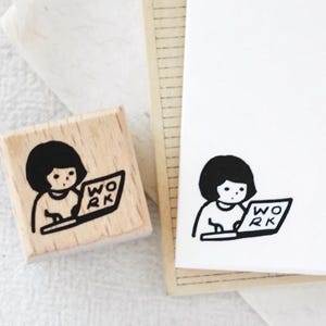 Puede incluir: Un sello de madera con una ilustraci&oacute;n en blanco y negro de una persona trabajando en un port&aacute;til. La palabra "WORK" est&aacute; en la pantalla. Una imagen estampada del mismo dise&ntilde;o est&aacute; en papel blanco.