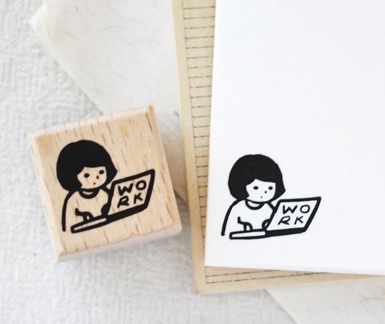 Puede incluir: Un sello de madera con una ilustraci&oacute;n en blanco y negro de una persona trabajando en un port&aacute;til. La palabra "WORK" est&aacute; en la pantalla. Una imagen estampada del mismo dise&ntilde;o est&aacute; en papel blanco.