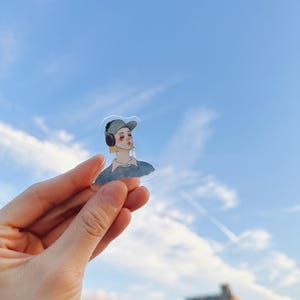 May include: A clear sticker featuring a watercolor illustration of a person wearing a cap and headphones. The person has blonde hair and rosy cheeks. The sticker is held up against a blue sky with clouds.