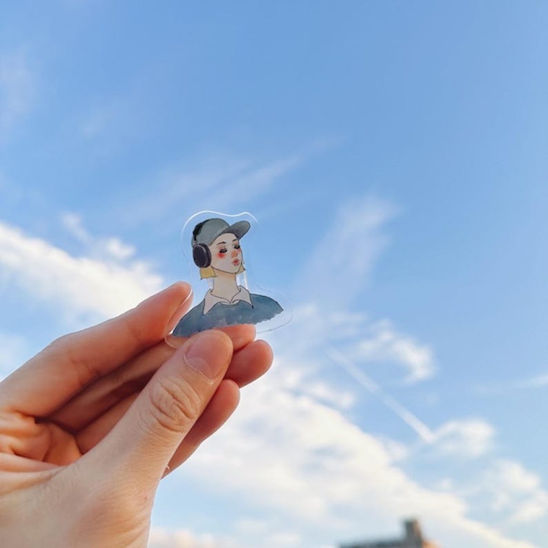 May include: A clear sticker featuring a watercolor illustration of a person wearing a cap and headphones. The person has blonde hair and rosy cheeks. The sticker is held up against a blue sky with clouds.