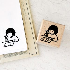 Puede incluir: Un sello de madera con una ilustraci&oacute;n negra de una persona escribiendo en un libro. El sello es cuadrado y tiene un acabado de madera clara. La ilustraci&oacute;n tambi&eacute;n est&aacute; impresa en papel blanco.