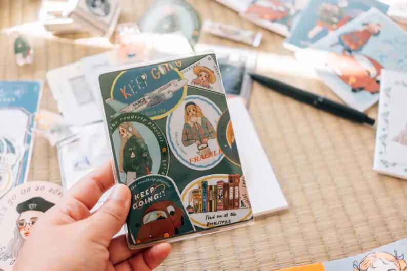 May include: A sheet of stickers with various illustrations and text. The stickers feature a plane, a book, a girl with a book, a girl with a backpack, and a car. The text on the stickers includes "Keep Going", "The Wanderlust Playlist", "Fragile", and "Find me at the Bookstore".