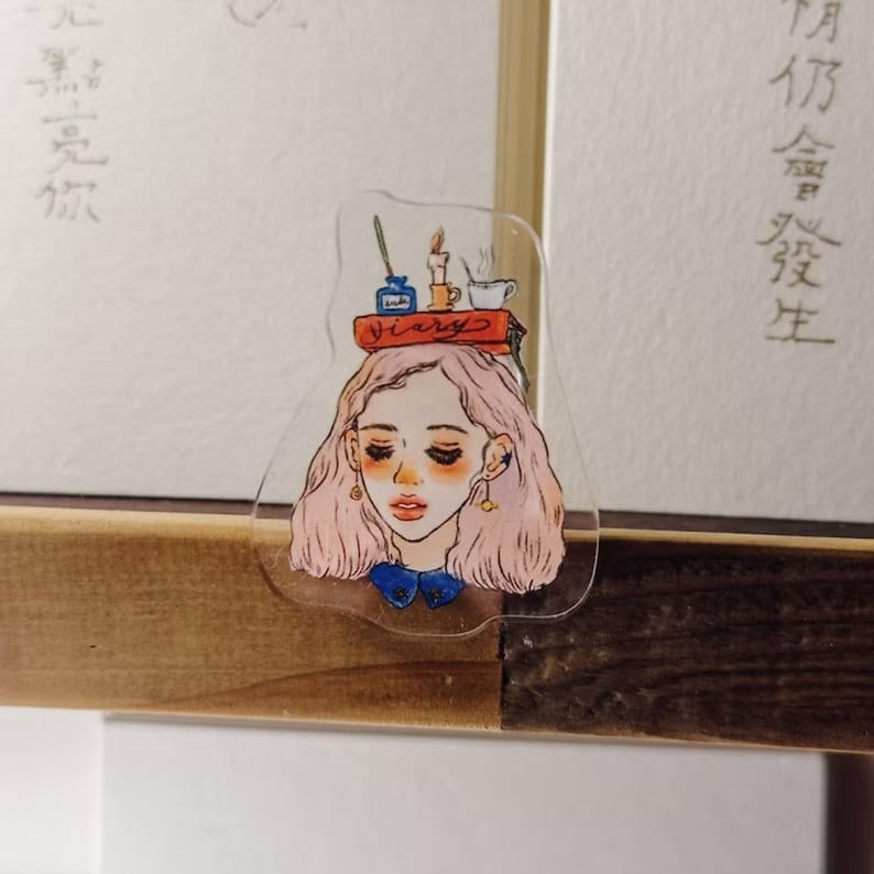 May include: A clear acrylic bookmark featuring a watercolor illustration of a woman with pink hair, wearing a blue bow. A stack of books with the word "Diary" is balanced on her head, along with a candle, inkwell, and teacup.