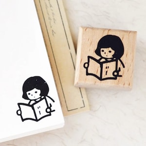 Puede incluir: Un sello de madera con una ilustraci&oacute;n en tinta negra de una persona leyendo un libro. El dise&ntilde;o es sencillo y de estilo caricaturesco, ideal para &aacute;lbumes de recortes o diarios.