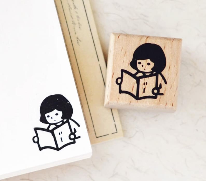 Puede incluir: Un sello de madera con una ilustraci&oacute;n en tinta negra de una persona leyendo un libro. El dise&ntilde;o es sencillo y de estilo caricaturesco, ideal para &aacute;lbumes de recortes o diarios.