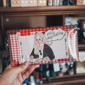 May include: A rectangular clear plastic pouch with a red and white gingham border containing stationery. The front features an illustration of a person writing in a notebook with the text Daily Inspiration in a speech bubble and small floral accents.