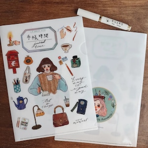 May include: Two clear plastic document folders with illustrations and text. One folder features a woman with a mug, the text "journal time", and other graphics. The other folder has a circular graphic and text. A white pen with gold accents rests on the folders.