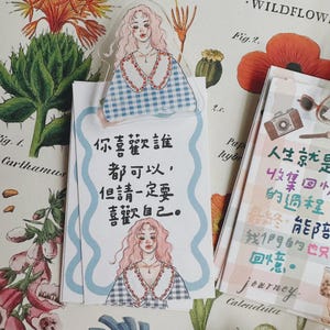 May include: Decorative paper items with illustrations and text. A clear bookmark features a girl illustration. A notepad has Chinese text and a girl illustration. Another notepad has Chinese text and a camera illustration. The items are on a floral background.