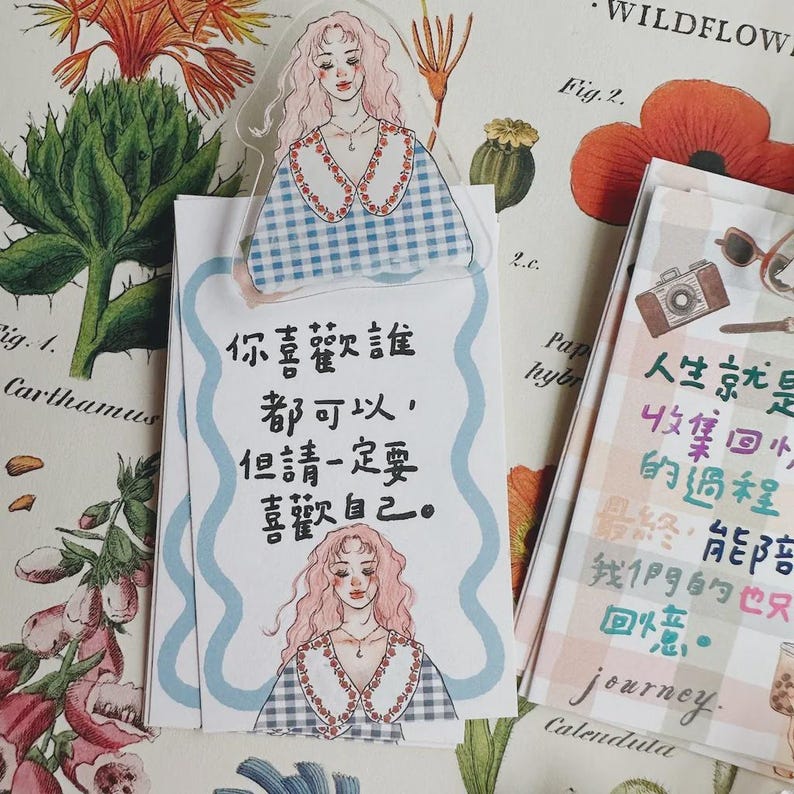 May include: Decorative paper items with illustrations and text. A clear bookmark features a girl illustration. A notepad has Chinese text and a girl illustration. Another notepad has Chinese text and a camera illustration. The items are on a floral background.