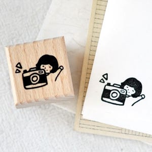 Puede incluir: Un sello de madera con una ilustraci&oacute;n en blanco y negro de una persona con una c&aacute;mara. El sello est&aacute; junto a un papel con la misma ilustraci&oacute;n. El dise&ntilde;o incluye formas geom&eacute;tricas.