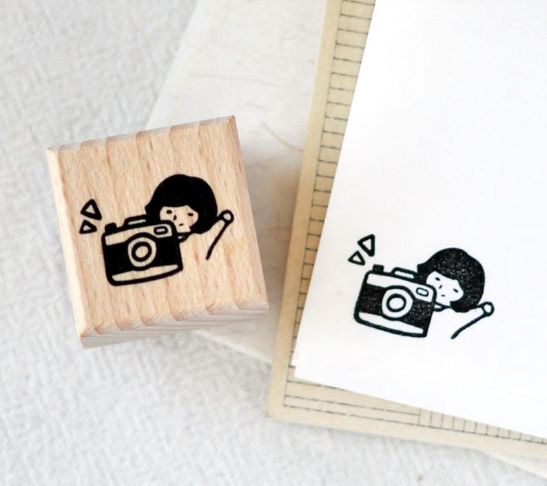 Puede incluir: Un sello de madera con una ilustraci&oacute;n en blanco y negro de una persona con una c&aacute;mara. El sello est&aacute; junto a un papel con la misma ilustraci&oacute;n. El dise&ntilde;o incluye formas geom&eacute;tricas.