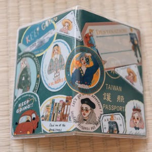 May include: A clear plastic passport holder with a green background and various colorful stickers. The stickers feature illustrations of people, airplanes, books, and text that says "Keep Going!" and "Let's go on an adventure!" The passport holder also has a sticker that says "Taiwan Passport" in English and Chinese.