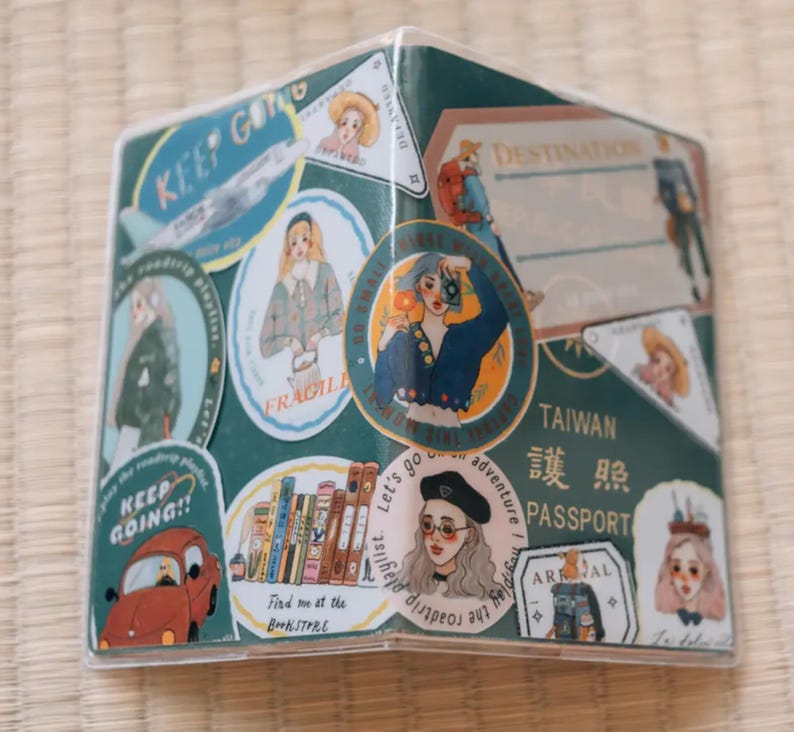 May include: A clear plastic passport holder with a green background and various colorful stickers. The stickers feature illustrations of people, airplanes, books, and text that says "Keep Going!" and "Let's go on an adventure!" The passport holder also has a sticker that says "Taiwan Passport" in English and Chinese.