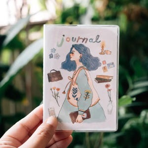 May include: A white journal cover with a blue-haired girl illustration. The girl is wearing a light blue dress with a floral pattern. The cover also features a variety of small illustrations, including flowers, a suitcase, books, and a candle.