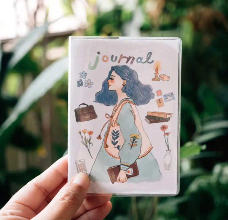 May include: A white journal cover with a blue-haired girl illustration. The girl is wearing a light blue dress with a floral pattern. The cover also features a variety of small illustrations, including flowers, a suitcase, books, and a candle.