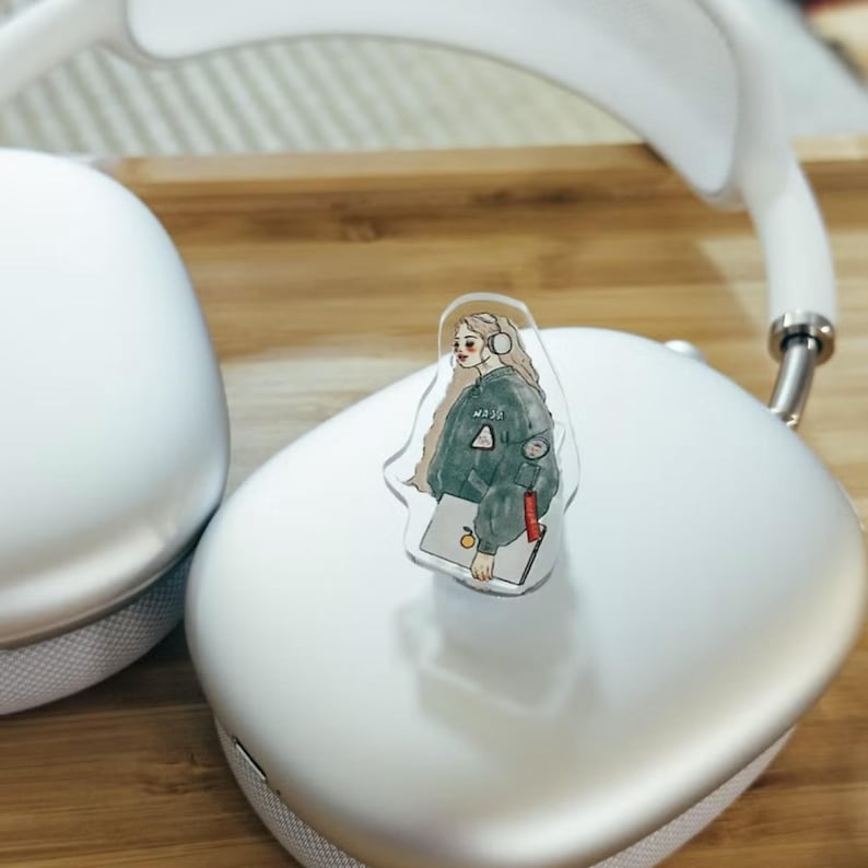May include: White over-ear headphones with a clear sticker featuring a cartoon illustration of a person wearing headphones and a green jacket. The sticker is placed on top of the right headphone cup. The background is a wooden surface.