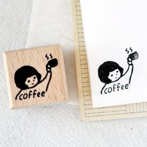 Puede incluir: Un sello de madera con una ilustraci&oacute;n negra de una persona sosteniendo una taza de caf&eacute;, con la palabra "coffee" debajo. Una imagen estampada a juego est&aacute; en papel blanco.