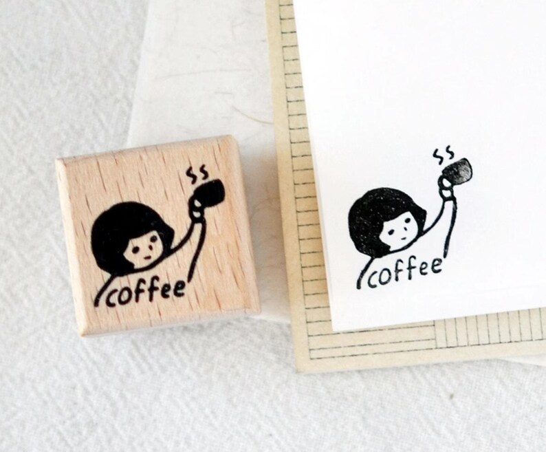Puede incluir: Un sello de madera con una ilustraci&oacute;n negra de una persona sosteniendo una taza de caf&eacute;, con la palabra "coffee" debajo. Una imagen estampada a juego est&aacute; en papel blanco.