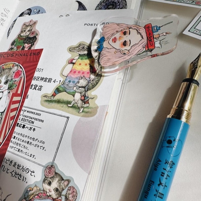 May include: A blue fountain pen with gold accents rests on a notebook with stickers of cats and a crocodile. A clear acrylic bookmark with a pink-haired woman illustration is on the notebook.