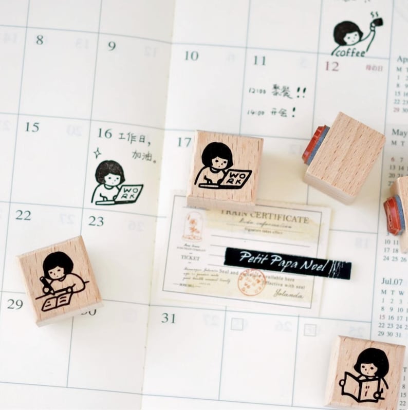 Puede incluir: Sellos de madera con ilustraciones de una ni&ntilde;a trabajando, leyendo y tomando caf&eacute;. Los sellos est&aacute;n en una p&aacute;gina de calendario con notas manuscritas y el texto "Petit Papa Noel".