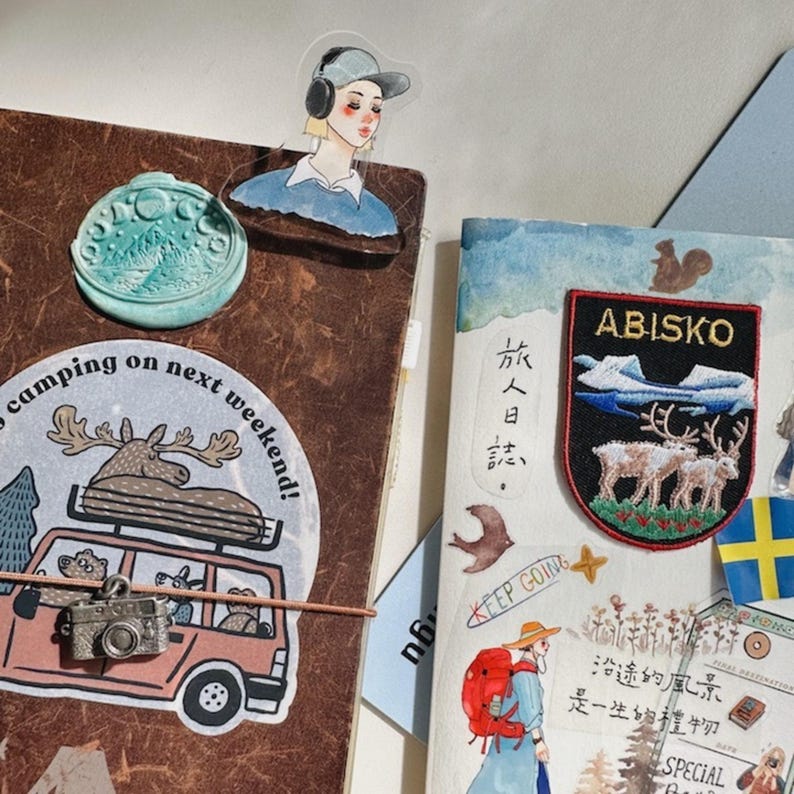 May include: A collection of decorative stickers and patches on a brown journal. Includes a sticker with a cartoon of a pink car, a camera, and the text "camping on next weekend!". Also features a patch with the text "ABISKO" and a drawing of reindeer.