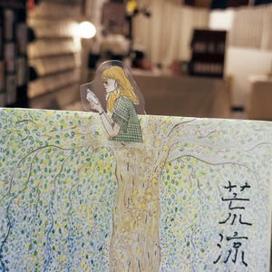 May include: A bookmark featuring an illustration of a woman reading, perched on a tree branch. The tree is painted with green and yellow leaves, and the bookmark is placed on a book with Japanese text. The woman has blonde hair and is wearing a green checkered shirt.
