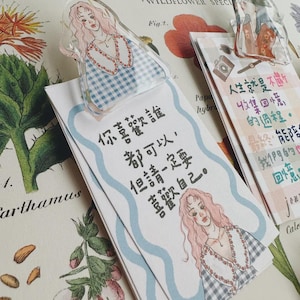May include: A clear acrylic bookmark with a pink-haired woman in a blue and white checkered dress, placed over a paper bookmark with Chinese text and a matching illustration. Botanical illustrations form the background.