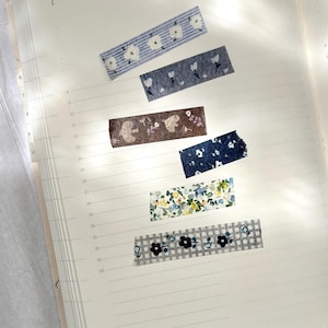 May include: An open notebook with several decorative washi tape strips. The tapes feature floral and patterned designs in various colours, including blue, brown, and green. The notebook pages have horizontal lines and numbered markings.