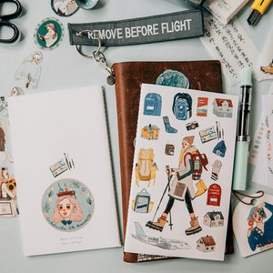 May include: A collection of stationery items, including stickers, notebooks, and a pen, arranged on a light blue surface. The stickers feature illustrations of people, houses, and travel-themed designs. A leather-bound journal and a "REMOVE BEFORE FLIGHT" tag are also visible.