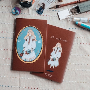 May include: Two brown notebooks with illustrations of a woman in a white dress holding flowers. One notebook has an oval frame with Chinese characters and the other says "La dolce vita". The notebooks are on a textured surface.