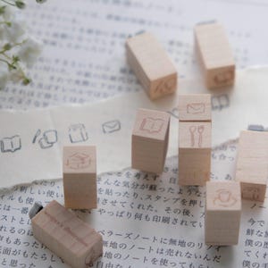 May include: A collection of small, rectangular wooden stamps featuring engraved icons such as an open book, a spoon and fork, a coffee cup, an envelope, and a package. The stamps are arranged on a page with Japanese text and small ink impressions.