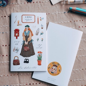 May include: A notebook with a decorative cover featuring a girl illustration, text in English and Japanese, and various stationery-themed graphics. The open notebook reveals blank pages. A sticker on the back cover reads "La dolce vita 10th Anniversary."