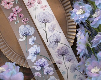Puede incluir: Dos rollos de cinta washi con diseños florales. La cinta de la izquierda tiene flores rosas y moradas, mientras que la cinta de la derecha tiene flores azules y moradas. Ambas cintas tienen un efecto translúcido, como de acuarela.