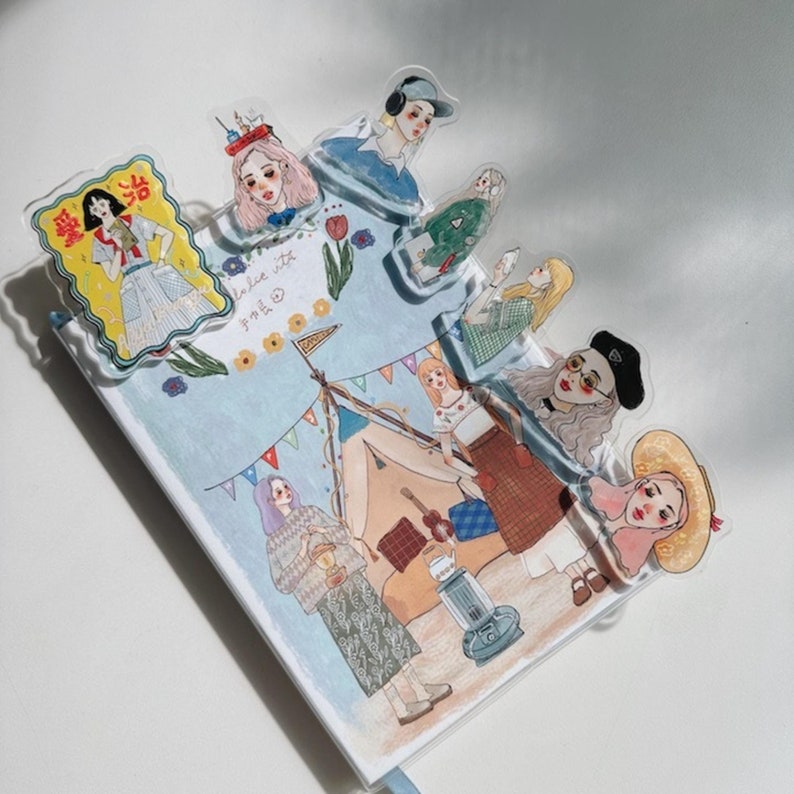 May include: A decorative journal with illustrations of women, a tent, and a guitar. Clear plastic bookmarks with illustrations of women are placed on the journal. The journal has the text "I'm here" and other text in a foreign language.