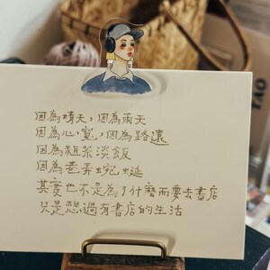 May include: A decorative bookmark featuring an illustration of a person wearing a cap and headphones. The bookmark is placed on a white card with Chinese text and a gold-colored metal clip.