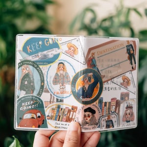 Può includere: Un foglio di plastica trasparente con adesivi colorati che mostrano illustrazioni di persone, testo e un aereo. Gli adesivi hanno frasi come "Continua", "Destinazione", "Andiamo all'avventura" e "Trova me in libreria".